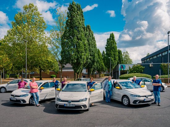 Eine Gruppe von Menschen posiert lächelnd neben mehreren weißen Autos auf einem Parkplatz. Im Hintergrund stehen hohe Bäume und ein großes Gebäude unter einem blauen Himmel mit weißen Wolken.