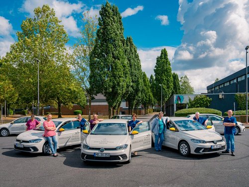 Eine Gruppe von Menschen posiert lächelnd neben mehreren weißen Autos auf einem Parkplatz. Im Hintergrund stehen hohe Bäume und ein großes Gebäude unter einem blauen Himmel mit weißen Wolken.